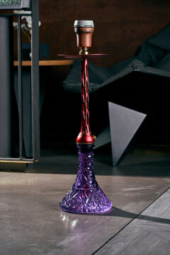 Modern purple glass hookah with red stem standing on floor in dark lounge interior.
