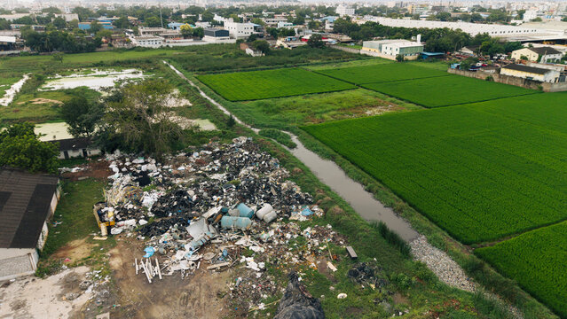 Aerial view of green farmland adjacent to a large waste dump, highlighting the contrast between agricultural land and environmental pollution in India.