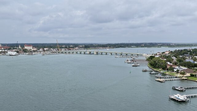 Aerial view of the Bridge of Lions spanning the Matanzas River with boats docked at piers and the historic St. Augustine skyline under a cloudy sky in St. Augustine, Florida, United States.