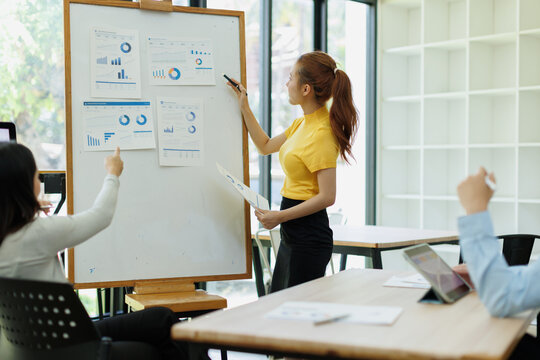 Businesswoman leading a corporate presentation, presenting financial data and analytics using charts on a whiteboard during an office meeting with colleagues discussing strategy