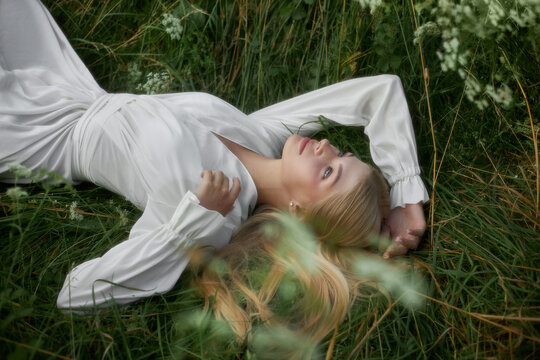 A woman lies on green grass in a field, wearing a white dress. She is gazing upwards with a thoughtful expression during daytime. The scene captures nature around her softly