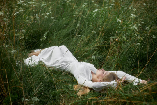 A woman lies on her back in a field of tall grass and wildflowers. She wears a white dress and enjoys the sunlight on a warm day. The sky is clear above her