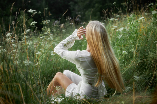 A woman sits on the ground in a green field with tall grass and wildflowers. She is looking upward, enjoying the clear sky and warm weather. Sunlight is shining through the trees nearby