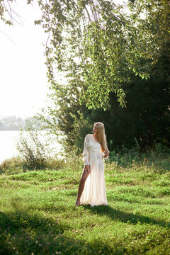 A woman is standing on green grass near a lake during the day. She wears a white dress and is surrounded by trees and sunlight. Water can be seen in the background