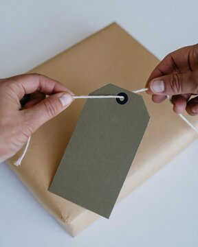 Hands threading a navy twine cord through a blank kraft tag on a wrapped parcel, featuring a tactile fibrous texture for stationery mockups