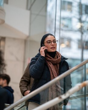 Middle Eastern woman in a hijab talking on a phone, standing by a ribbed glass rail in a bright modern office for business and corporate use