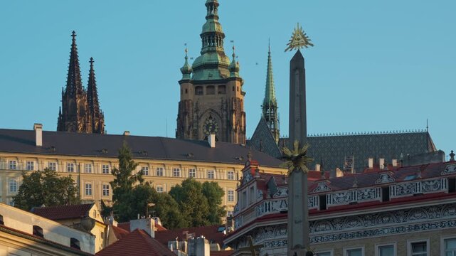 Holy Trinity Column stands on Malostranske Square in Prague. Historic spires of Saint Vitus Cathedral and Prague Castle rise in the background. European urban architecture in morning light.
