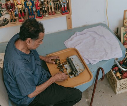 Antique wooden toys and vintage collectibles on a shelf behind a man repairing a weathered wind-up mechanism for collector media and hobbies