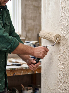 Man applying textured plaster with a roller to an interior wall. Professional renovation work creates a decorative textured finish