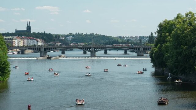 Tourists paddle pedal boats on the Vltava River near historic Palacky Bridge in Prague. A river weir cascades water near Vysehrad castle under a bright summer sky. Popular water recreation.