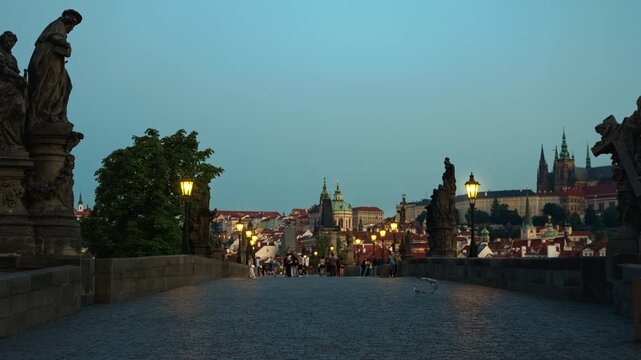 Tourists walk on historic Charles Bridge in Prague at dusk with glowing lanterns. Famous Czech capital landmark with cobblestones and castle view. Ideal for European travel and tourism content.