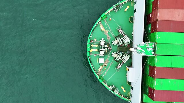 multiple ship crew members in bright safety orange working with massive ropes and heavy machinery on the vibrant green deck of a large vessel's bow, with blue water surrounding
