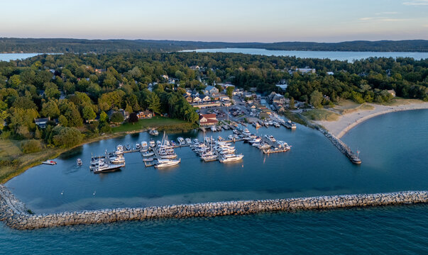 Aerial view of Leland marina with boats docked in the calm harbor, protected by a stone breakwater, surrounded by lush green trees and a sandy beach in Leland, Michigan, United States.