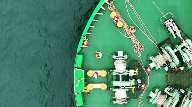 multiple ship crew members in bright safety orange working with massive ropes and heavy machinery on the vibrant green deck of a large vessel's bow, with blue water surrounding