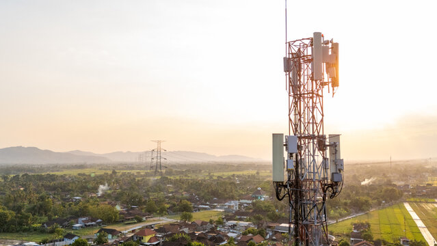 Aerial view of a telecommunications tower standing above a rural landscape with houses, green fields, and distant mountains under a warm golden hour sky in Klaten, Central Java, Indonesia.