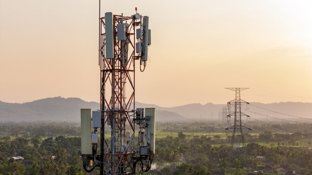 Aerial view of a telecommunications tower amidst rural greenery and power lines with distant mountains under a golden hour sky in Klaten, Central Java, Indonesia.