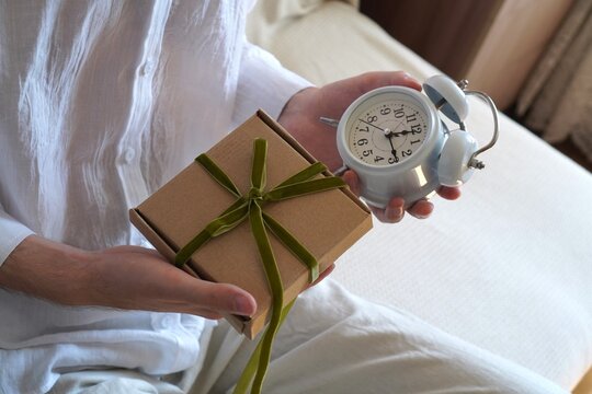 A man holding a craft gift box with a green ribbon and an alarm clock. On-time express gift delivery service concept for holidays, Christmas, and birthdays. Fast shipping and deadline concept