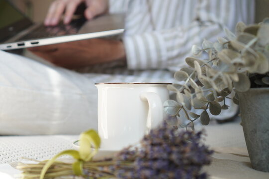 White mug, lavender, and eucalyptus in the foreground with a man typing on a laptop blurred behind. Cozy domestic workplace for remote work, freelancing, digital business, and online study