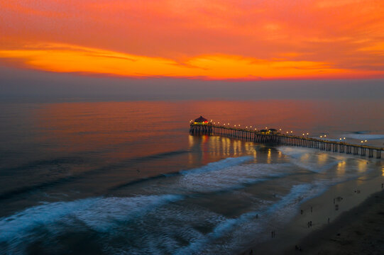 Aerial view of Huntington Beach Pier at sunset, with vibrant orange sky, gentle waves, and illuminated pier lights in Huntington Beach, California, United States.