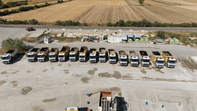 Aerial view of a row of dump trucks parked in a gravel lot with agricultural fields in the background in Kuzmin, Vojvodina, Serbia.