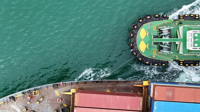 multiple ship crew members in bright safety orange working with massive ropes and heavy machinery on the vibrant green deck of a large vessel's bow, with blue water surrounding