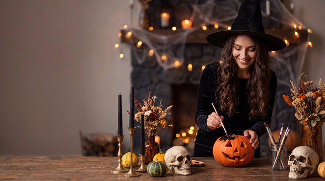 Woman in witch hat painting a Halloween pumpkin on a wooden table, with decorative skulls and autumn-themed elements in a cozy setting