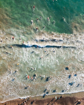 Aerial view of surfers waiting for waves in the turquoise ocean with surfboards and white foam along the sandy beach in Orange County, California, United States.
