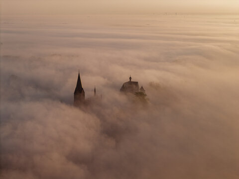 Aerial view of Keverberg Castle emerging from a thick layer of morning fog, with the church spire and castle towers visible above the mist in Kessel, Limburg, Netherlands.