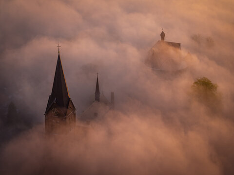 Aerial view of church spires rising above a thick layer of golden fog at sunrise in Kessel, Limburg, Netherlands.