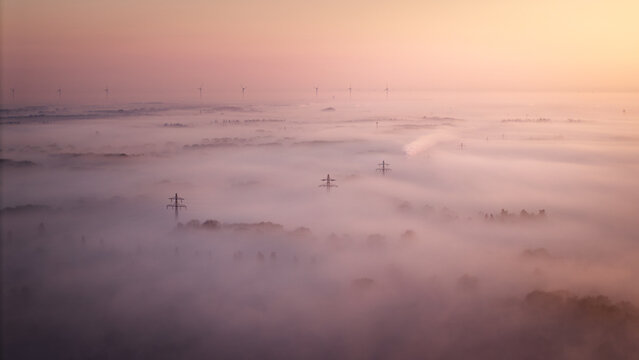 Aerial view of a landscape shrouded in thick morning fog with electricity pylons rising above the mist and wind turbines visible on the horizon in Kessel, Limburg, Netherlands.