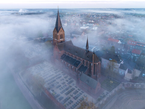 Aerial view of the Church of the Nativity of Our Lady surrounded by a cemetery and the foggy village of Kessel, Limburg, Netherlands.
