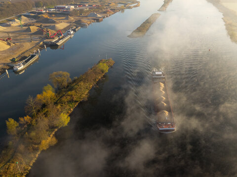 Aerial view of a cargo barge transporting sand along a river in the early morning mist, passing an industrial site with stockpiles of gravel in Kessel, Limburg, Netherlands.