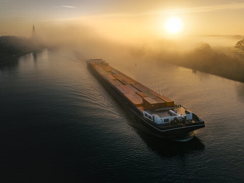 Aerial view of a cargo barge navigating a river during golden hour, surrounded by morning fog and calm water in Kessel, Limburg, Netherlands.