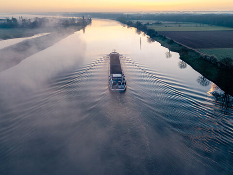 Aerial view of a cargo barge navigating a calm river during a misty sunrise with golden light reflecting on the water surface in Kessel, Limburg, Netherlands.