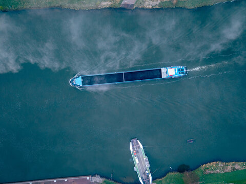 Aerial view of a cargo barge navigating a river with a ferry docked at the bank in the misty morning light in Kessel, Limburg, Netherlands.