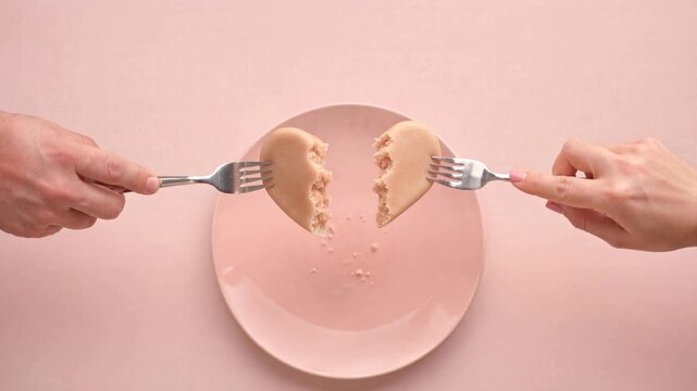 Hands breaking a heart-shaped cookie with forks on a pink plate, dessert close-up symbolizing division, conflict, heartbreak and end of love with playful pastel styling
