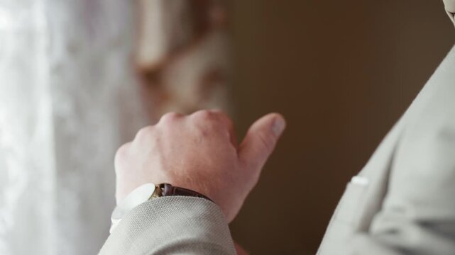 A groom stands near a window, checking his watch while waiting for the wedding ceremony to start. The light shines through, creating a warm atmosphere.