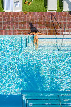 Aerial view of a woman relaxing in a swimming pool with clear blue water, surrounded by a brick deck and lounge chairs in Newport Beach, California, United States.