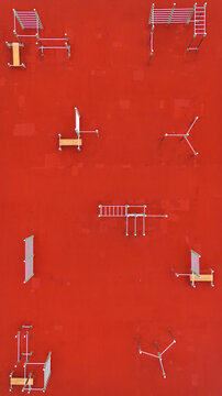 Aerial view of a modern outdoor calisthenics park with metal exercise equipment and benches set against a vibrant red rubberized ground surface in Riga, Latvia.