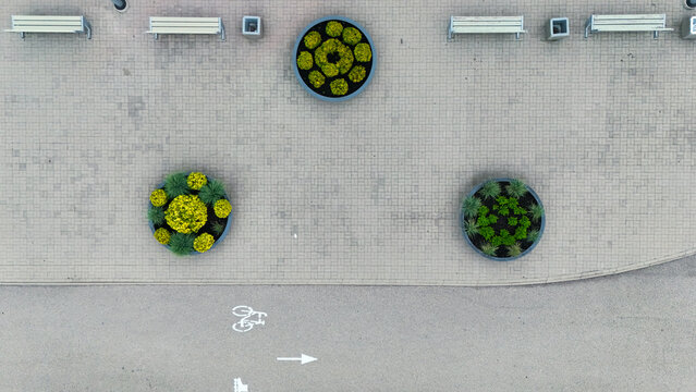Aerial view of circular planters with greenery and benches on a paved walkway next to a bicycle lane in Riga, Latvia.
