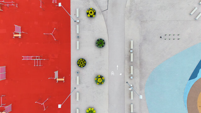 Aerial view of a modern urban playground with a vibrant red surface, circular planters along a paved walkway, and geometric patterns in Riga, Latvia.