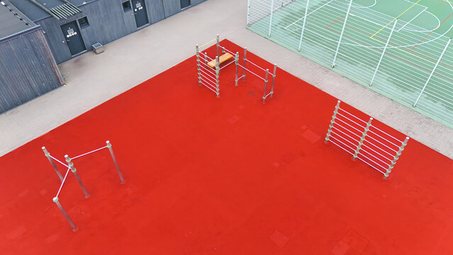 Aerial view of an outdoor fitness area with red rubber flooring, metal pull-up bars, and exercise equipment located next to a sports court in Riga, Latvia.