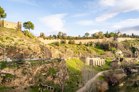 View of the historic fortified walls of Toledo overlooking the Tagus River with rocky terrain and greenery under a clear blue sky in Toledo, Castile-La Mancha, Spain.