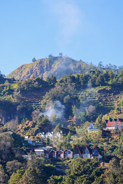 Cozy village nestles into green hillsides at Dieng Wonosobo. Smoke curls gently from distant terraced fields. Sunlit rooftops contrast with deep forested slopes. Climbing hillsides hint at ancient cul