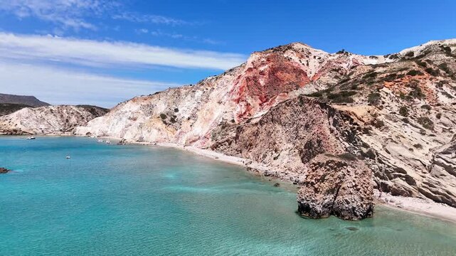 Aerial view of Paralia Firiplaka on Milos, Greece, showing clear turquoise water, a narrow sandy beach, layered volcanic cliffs, and a small red boat near shore.