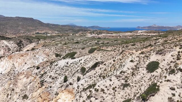 Aerial footage above Paralia Firiplaka on Milos, Greece, reveals pale eroded ravines, sparse shrubs, rolling volcanic terrain, and a strip of deep blue Aegean Sea in the distance.