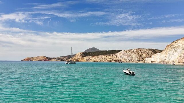 Wide coastal view at Paralia Firiplaka on Milos, Greece, shows a small motorboat and sailboat on turquoise water beneath pale volcanic cliffs and a streaked summer sky.
