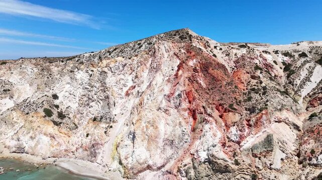 Detailed aerial view of layered white, red, and gray volcanic cliffs at Paralia Firiplaka on Milos, Greece, with sparse shrubs scattered across the rugged slope.