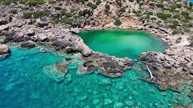 Aerial view of Voulolimni in Crete, Greece, showing an emerald natural pool enclosed by pale rocks beside clear blue sea, submerged stones, and sparse coastal shrubs.