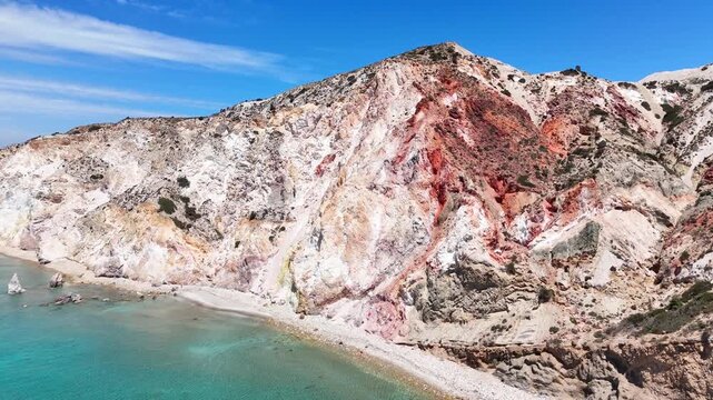 Aerial footage of Paralia Firiplaka on Milos, Greece, where shallow blue water meets a quiet curved beach below striking white and rust colored volcanic cliffs.